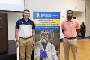 Two men stand beside a North Carolina A&T State University sign for the Undergraduate Research and Creativity Symposium in a large indoor space. One wears a white polo and khakis, the other a salmon polo and khakis.