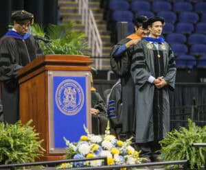 A graduate in academic regalia stands smiling on stage as another person places a hood over his shoulders during a graduation ceremony. A speaker stands at a podium adorned with a university seal and a floral arrangement.