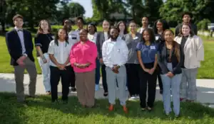 A group of 18 people, dressed in business casual attire, stand outdoors on grass and a sidewalk, smiling at the camera on a sunny day with trees and buildings in the background.