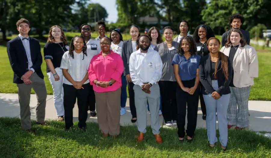 A group of 18 people, dressed in business casual attire, stand outdoors on grass and a sidewalk, smiling at the camera on a sunny day with trees and buildings in the background.