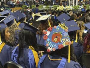 Graduates in blue caps and gowns sit at a commencement ceremony. One cap is decorated with a red apple, a school bus, and the phrase “My Turn to Teach,” featuring colorful school-themed embellishments.