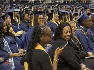 Rows of graduates in navy blue caps and gowns sit in an auditorium. Some smile and look at the camera, while others face forward. A woman in the front row holds a phone and another smiles beside her.