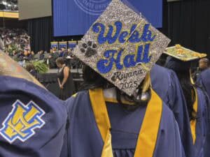 A graduate in a navy and gold gown stands at a ceremony, wearing a decorated cap that reads Welk Hall, Room 216, 2025 with paw prints and rhinestones. Other graduates are visible in the background.
