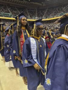 A group of graduates in blue gowns and caps stand in line at a crowded indoor graduation ceremony. One holds a diploma cover, and the arena is filled with spectators.