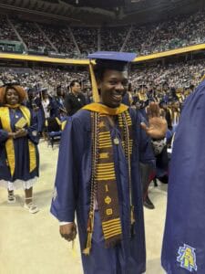 A smiling graduate in a blue cap and gown waves at the camera during a graduation ceremony in a crowded indoor arena, with other graduates and attendees in the background.