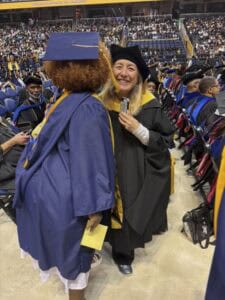 A graduate in a blue cap and gown hugs a smiling faculty member in black academic regalia during a commencement ceremony, with rows of seated graduates and a cheering audience in the background.