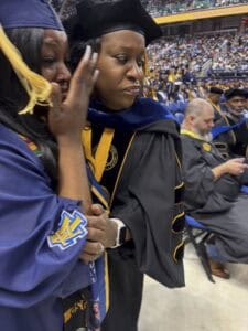 A graduate in a navy cap and gown wipes away tears while being comforted by a faculty member in academic regalia during a graduation ceremony in a crowded arena.