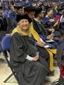 A woman in academic regalia sits and smiles at a graduation ceremony, surrounded by other faculty members in similar robes and caps, with a crowd and graduates in the background.