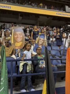 A crowd at a graduation ceremony holds large cutouts of a graduate in a cap and gown, with more people standing and watching from the stadium seats behind them.