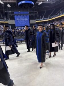 Graduates in academic regalia smile and walk inside an arena during a university commencement ceremony. Other graduates and attendees fill the background, with a large blue stage and banners visible.