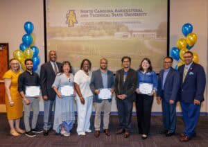 A group of ten people poses indoors in front of a screen displaying North Carolina Agricultural and Technical State University. Five people in the center hold certificates; blue and gold balloons are on each side.