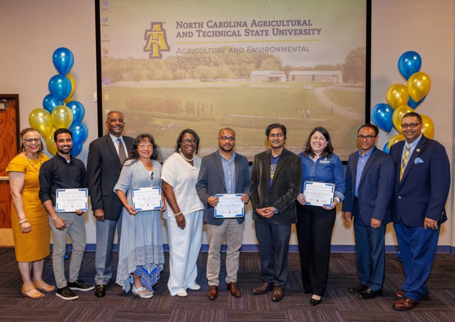 A group of ten people poses indoors in front of a screen displaying North Carolina Agricultural and Technical State University. Five people in the center hold certificates; blue and gold balloons are on each side.