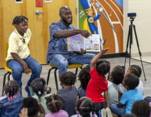 A man sits next to a boy, reading a picture book to a group of young children seated on the floor. One child has their hand raised. A mural and a camera on a tripod are visible in the background.