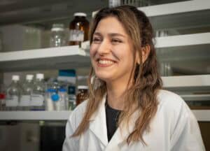 A smiling woman wearing a white lab coat stands in a laboratory with shelves of bottles and lab equipment in the background.