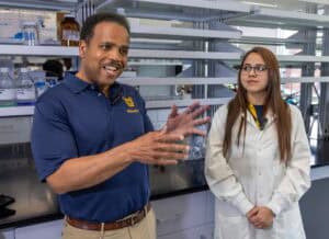 A man in a navy polo shirt gestures while speaking to a woman in a white lab coat standing beside him in a laboratory setting with shelves and scientific equipment in the background.