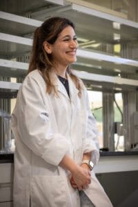 A woman wearing a white lab coat stands in a laboratory, smiling and looking off to the side. She has long brown hair and wears a watch and necklace. Laboratory shelves and equipment are visible in the background.