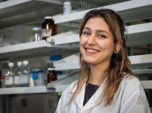 A young woman wearing a white lab coat smiles while standing in a laboratory with shelves containing bottles and glass containers in the background.