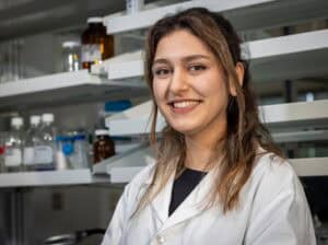 A young woman wearing a white lab coat smiles while standing in a laboratory, with shelves of glass bottles and scientific equipment in the background.