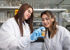 Two women in lab coats examine a beaker with an orange liquid in a laboratory setting. One woman is holding the beaker with gloved hands while they both smile and observe the contents closely.