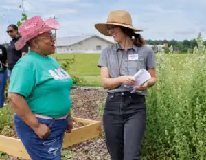Two women stand and chat in a community garden. One wears a pink cowboy hat and green t-shirt, the other wears a wide-brimmed straw hat, glasses, and holds papers. Raised garden beds and greenery appear in the background.