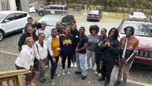 A group of people pose outdoors, smiling at the camera. They stand in a gravel parking area with parked cars behind them and trees in the background. Most are dressed casually in sweatshirts and athletic wear.