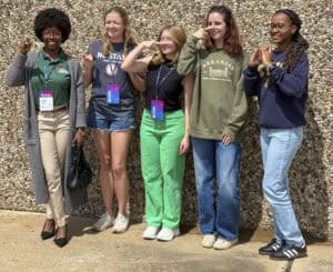 Five young women stand smiling in a row outside against a textured stone wall, each making a hand gesture with their right hand. They wear casual clothes and have event badges around their necks.