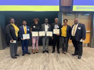 Seven people stand side by side indoors, five holding certificates. They are smiling and dressed in business or business casual attire. A sign above the door reads North Carolina Agricultural and Technical State University.