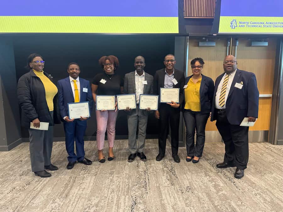 Seven people stand side by side indoors, five holding certificates. They are smiling and dressed in business or business casual attire. A sign above the door reads North Carolina Agricultural and Technical State University.