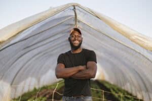 A man wearing a black t-shirt and orange cap stands smiling with arms crossed in front of a greenhouse structure made of transparent plastic. Green plants grow inside the greenhouse. The sky is clear.
