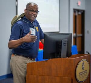 A man wearing glasses and a navy polo shirt speaks at a wooden podium with a computer monitor, in a classroom or lecture room at North Carolina A&T State University.