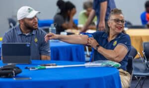 A woman in a navy polo shirt smiles while rolling up her sleeve at a table, next to a man wearing a white cap and polo shirt. Other people sit and talk at tables in the background.