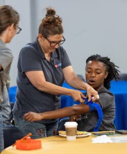A woman wraps a blue band around another woman’s arm as they sit at a table. The woman being helped looks surprised, and there’s food, a coffee cup, and other items on the table. Another person stands nearby.