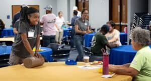 A woman leans on a table and smiles while talking to an older person seated across from her. Other people in casual clothing chat and move around in a brightly lit room with round tables covered in yellow and blue tablecloths.