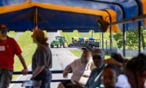 People ride in a covered wagon, looking ahead at another tractor pulling a similar passenger wagon down a rural road with green grass and trees on a sunny day.
