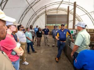 A group of people listen to a man in a navy shirt and cap speaking inside a large, white, open-ended tent structure. The group appears engaged, standing around him in a semi-circle.