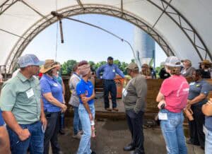A group of people stands in a covered outdoor structure, listening to a person in a blue shirt speak. Some wear hats and name tags. A metal silo and trees are visible in the background.