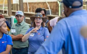 A woman in a blue NC State shirt and hat speaks passionately to a group of people indoors, using hand gestures. Several people listen attentively, some with arms crossed, all wearing name badges.