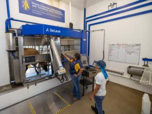 Two people observe a cow in a robotic milking machine in a modern dairy facility at North Carolina A&T State University. One person points at the machine while the other looks on. A whiteboard and sink are visible on the wall.