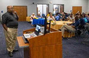 A man speaks into a microphone at a podium in a conference room, addressing an audience seated at round tables, some with laptops open and others listening attentively.