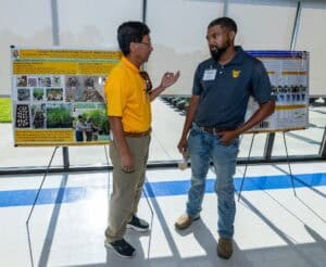 Two men stand and talk in front of scientific posters displayed on easels in a bright, modern indoor space. One man gestures as he speaks, while the other listens, holding a drink container.