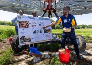 A man points to a research poster on animal feed substrates while another man holds a bucket of wood chips under a large irrigation system in a sunny field. A third man observes in the background.