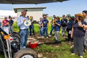 A man in a hat demonstrates equipment to a diverse group of adults outdoors on a sunny day. The group listens attentively, some taking notes or photos, surrounded by grass and farm buildings.