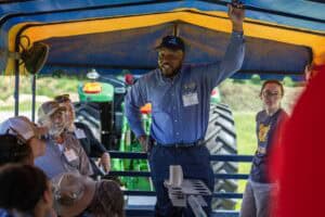 A man stands and speaks to a seated group under a blue and yellow canopy near a green tractor, while a woman and others listen attentively during a daytime outdoor event.