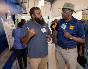 Two men engaged in conversation at an indoor event; one gestures with his thumb, the other wears a hat and holds his glasses. Informational posters and other attendees are visible in the background.