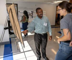 A man in a green shirt presents a research poster to a woman in glasses at a conference, while another person looks at posters in the background inside a brightly lit room.
