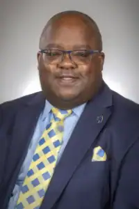 A man wearing glasses, a navy suit, a light blue shirt, and a yellow and blue patterned tie smiles at the camera against a neutral background.