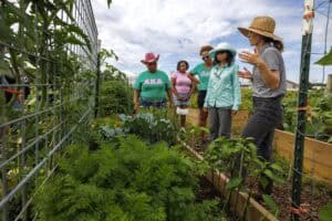 A group of five women stand together in a lush community garden, listening to one woman in a straw hat as she gestures and explains something about the plants growing in raised beds.