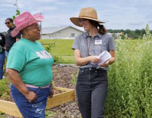 Two women talk and smile in a garden. One wears a pink cowboy hat and teal shirt, the other a wide-brimmed hat and gray shirt, holding papers. Raised garden beds and greenery are visible in the background.