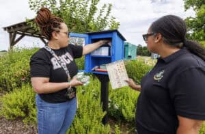 Two women are standing outside by a blue community pantry box. One places items inside while the other holds a chart. They are surrounded by greenery and garden plants under a partly cloudy sky.