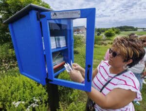 A woman takes a packet from a blue outdoor Little Free Library box filled with seed packets, surrounded by greenery and flowers on a sunny day.